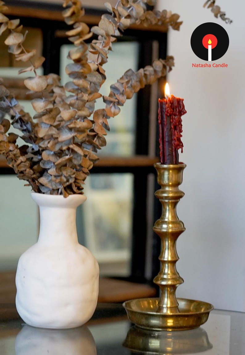May include: A lit dark red taper candle with visible wax drips sits in a polished brass candlestick holder. Next to it is a white ceramic vase containing dried eucalyptus branches. The logo Natasha Candle is visible in the upper right corner.