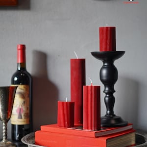 Peut inclure: Composition décorative avec des bougies rouges à piliers de différentes hauteurs, posées sur des livres rouges et un bougeoir noir. Une bouteille de vin rouge et un gobelet en argent sont également présents. Les bougies ont une texture côtelée.