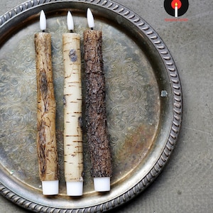May include: Three textured, battery-operated candles with white tips and bases, resembling tree bark. The candles are arranged on a silver, ornate tray. The candle on the left is a light brown color, the middle candle is off-white, and the candle on the right is dark brown.