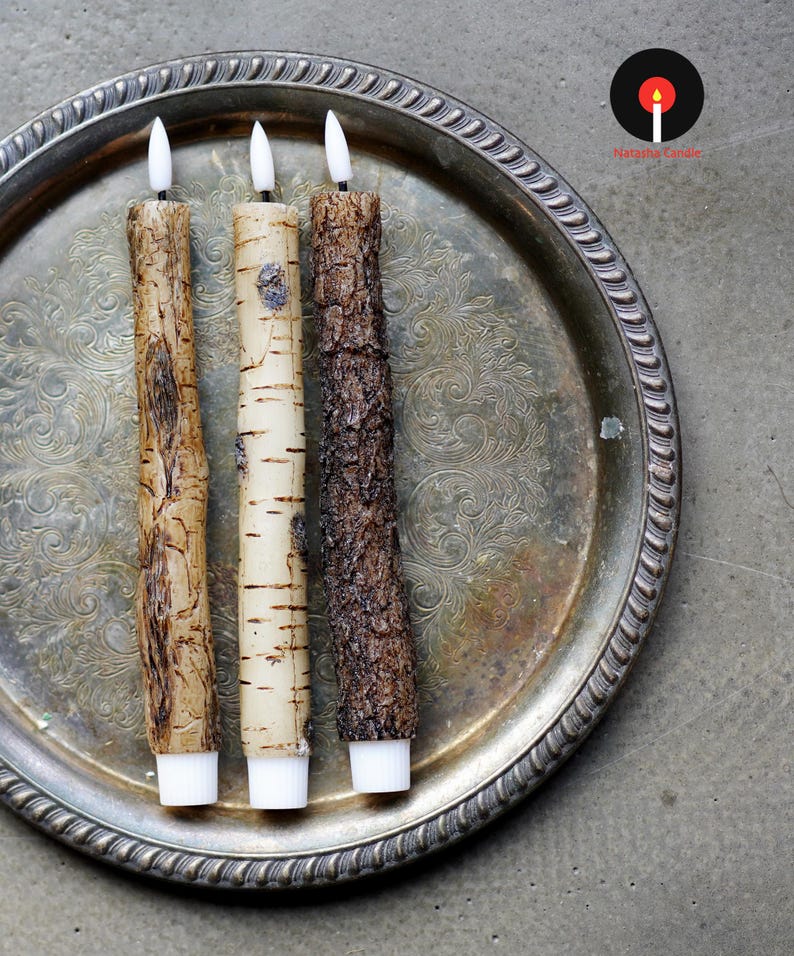 May include: Three textured, battery-operated candles with white tips and bases, resembling tree bark. The candles are arranged on a silver, ornate tray. The candle on the left is a light brown color, the middle candle is off-white, and the candle on the right is dark brown.