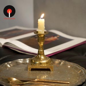 May include: A brass candlestick holder with a square base sits on a round, textured metal tray. A lit, off-white taper candle is inserted into the holder. A metal spoon and knife rest on the tray. The logo Natasha Candle is visible.