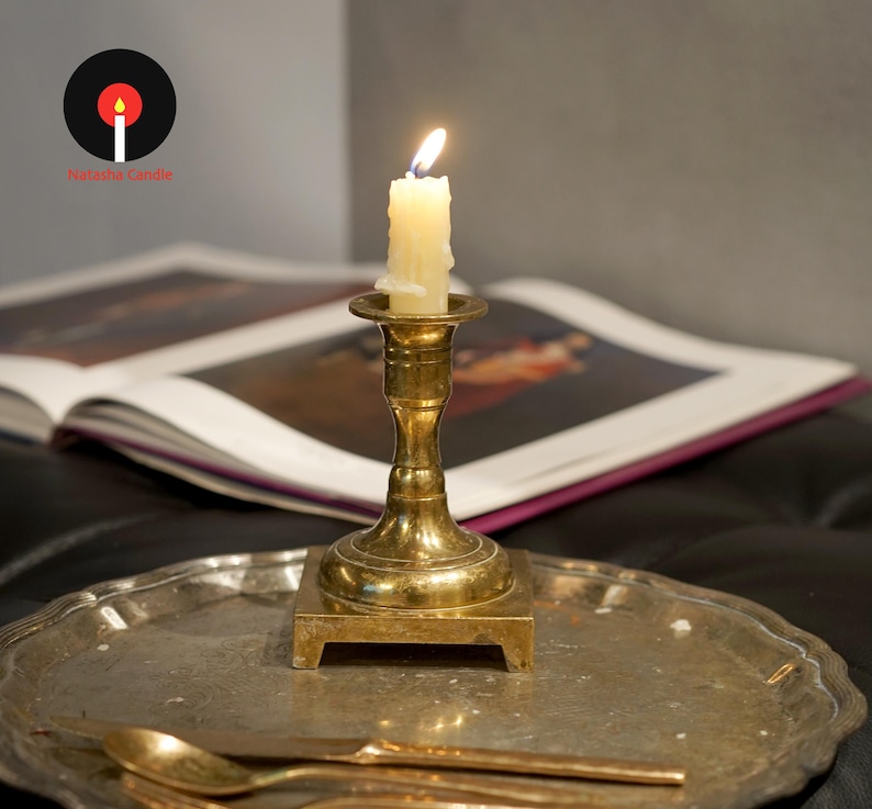 May include: A brass candlestick holder with a square base sits on a round, textured metal tray. A lit, off-white taper candle is inserted into the holder. A metal spoon and knife rest on the tray. The logo Natasha Candle is visible.