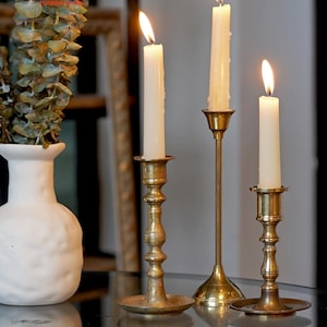 May include: Three brass candlesticks of varying heights, each featuring a slender stem and a circular base, hold lit white taper candles. The candlesticks are arranged in a row, with a white textured vase containing eucalyptus leaves nearby.
