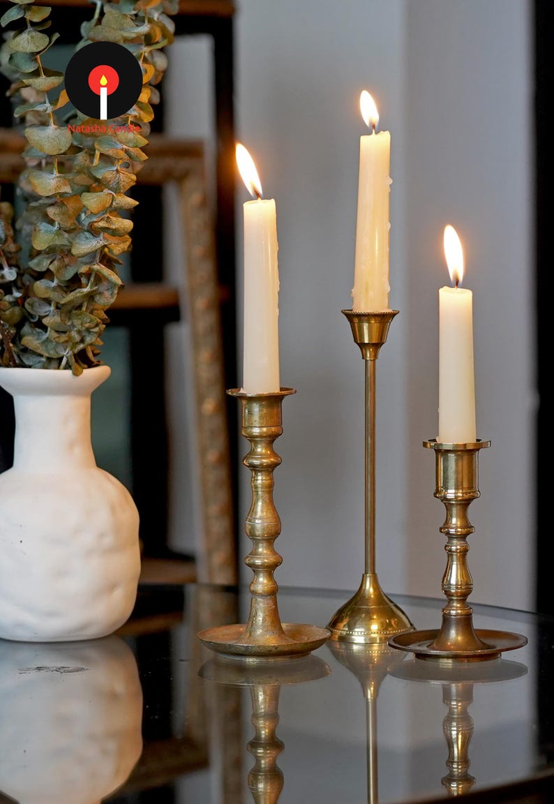May include: Three brass candlesticks of varying heights, each featuring a slender stem and a circular base, hold lit white taper candles. The candlesticks are arranged in a row, with a white textured vase containing eucalyptus leaves nearby.