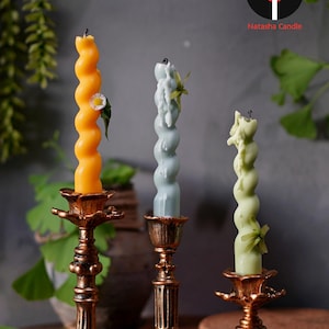May include: Three spiral-shaped candles in orange, blue-grey, and green, each adorned with a small flower or greenery. The candles are displayed in ornate, antique-style gold candle holders. The image includes the text "Natasha Candle".