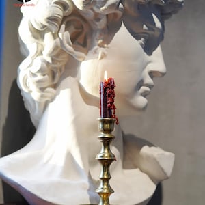 May include: A white plaster bust of a classical figure positioned behind a brass candlestick holder. A dark red candle with visible wax drips and a lit flame sits in the holder. The logo for Nasama Candle is visible in the upper left corner.