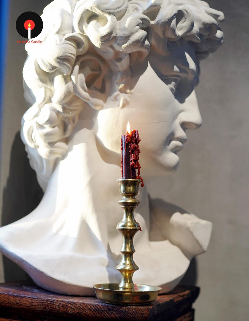 May include: A white plaster bust of a classical figure positioned behind a brass candlestick holder. A dark red candle with visible wax drips and a lit flame sits in the holder. The logo for Nasama Candle is visible in the upper left corner.