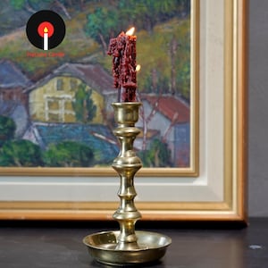 May include: A tall, polished brass candlestick holder with a tiered, cylindrical stem and a wide, circular drip tray base. A single, thick, dark red candle with visible wax drips and a lit flame sits securely in the top socket of the holder.