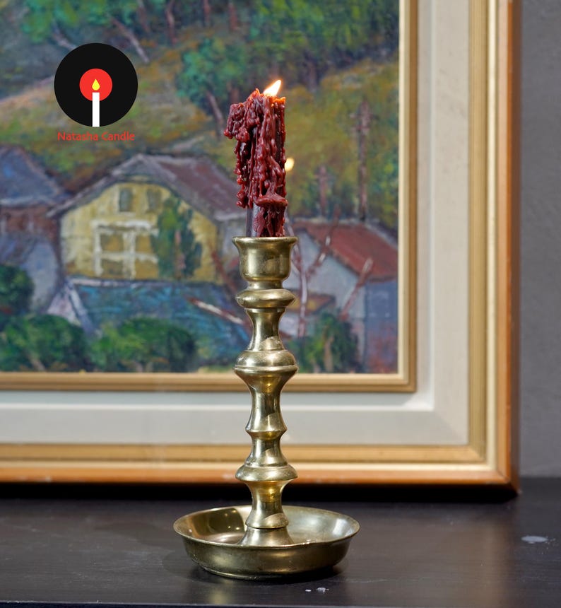 May include: A tall, polished brass candlestick holder with a tiered, cylindrical stem and a wide, circular drip tray base. A single, thick, dark red candle with visible wax drips and a lit flame sits securely in the top socket of the holder.