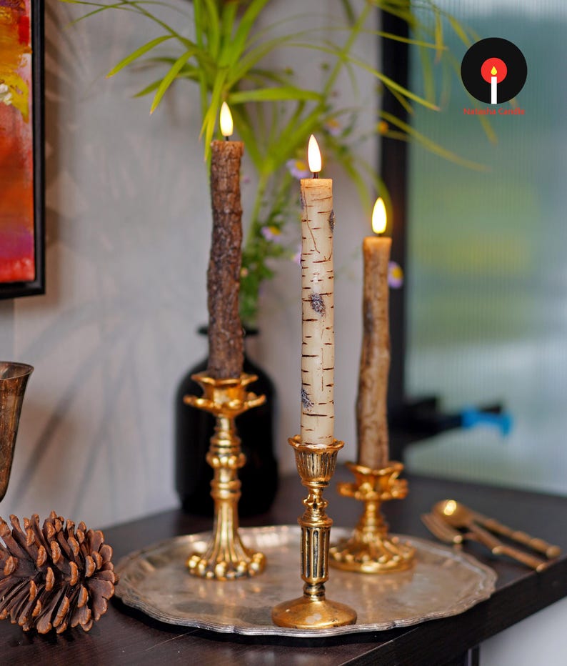 May include: Three lit candles in gold-colored ornate candle holders. The candles have a natural, textured appearance, with one resembling birch bark. The candles are on a silver tray, with a pinecone and decorative items in the background.
