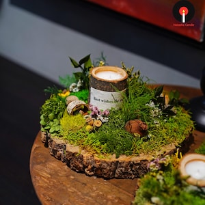 Puede incluir: Una vela decorativa en un soporte de madera, rodeada de musgo verde, pequeñas flores y vegetación. La vela tiene una etiqueta blanca que dice "Best wishes for you". La vela se encuentra sobre una base de rodaja de madera.