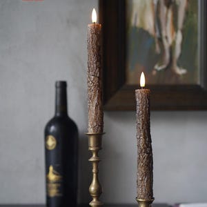 May include: Two brown textured candles with flickering flames sit in antique brass candle holders. A dark wine bottle is in the background. The candles have a rustic, bark-like appearance, creating a warm, inviting ambiance.