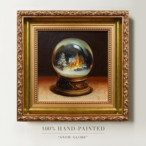 May include: A hand-painted framed artwork of a snow globe. The snow globe features a winter scene with a church and falling snow. The frame is gold with ornate detailing. The text at the bottom reads "100% HAND-PAINTED" and "SNOW GLOBE".