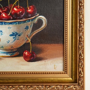 Puede incluir: Pintura enmarcada de naturaleza muerta con una taza de t&eacute; blanca y azul con estampado floral rebosante de cerezas rojas maduras. Una sola cereza descansa sobre la mesa. El marco dorado adornado rodea la obra de arte.