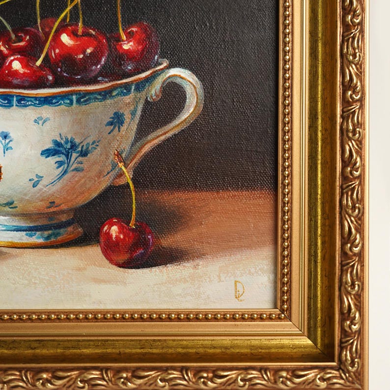Puede incluir: Pintura enmarcada de naturaleza muerta con una taza de t&eacute; blanca y azul con estampado floral rebosante de cerezas rojas maduras. Una sola cereza descansa sobre la mesa. El marco dorado adornado rodea la obra de arte.