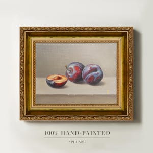 May include: An oil painting of two whole plums and one halved plum resting on a flat surface, set within an ornate, antique-style gold frame. The artwork features realistic shading and deep purple and red tones against a neutral background.