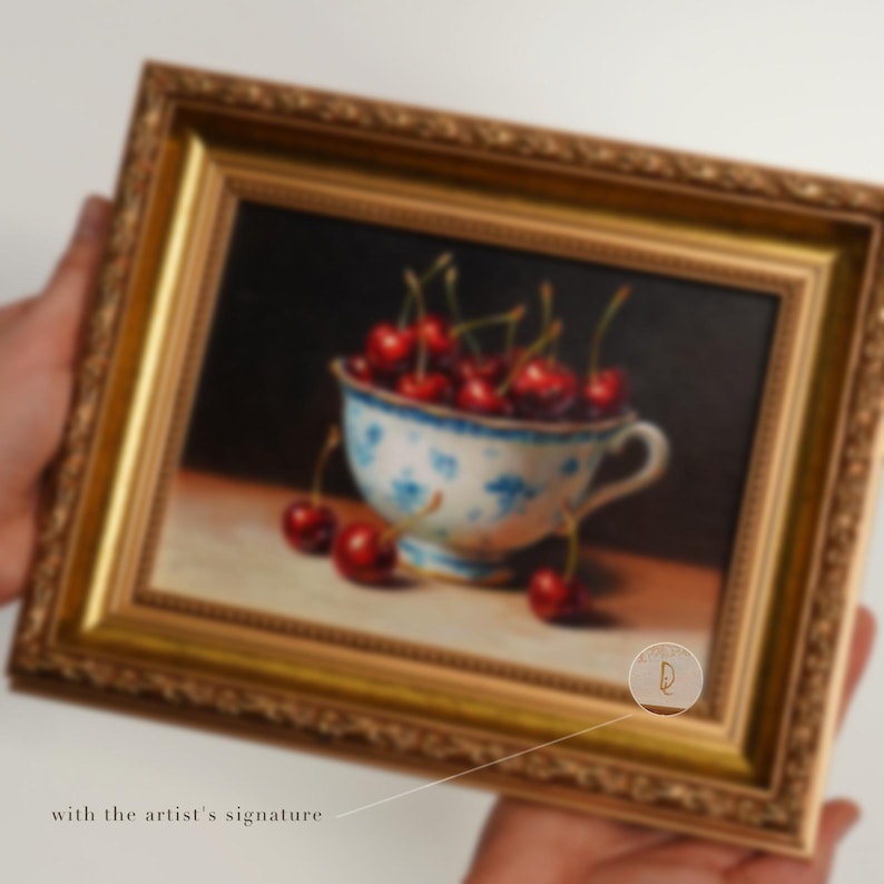 Puede incluir: Pintura enmarcada de naturaleza muerta con una taza llena de cerezas maduras. La taza tiene un dise&ntilde;o floral azul y blanco. La pintura est&aacute; en un marco dorado y tiene la firma del artista.