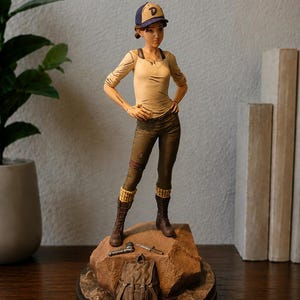 Op de afbeelding: Een verzamelfiguur van een jong persoon met een blauwe en beige honkbalpet met de letter D, een beige shirt met lange mouwen en een donkere broek. Het figuur staat op een rotsachtige basis met een kleine rugzak, een mes en een pistool.