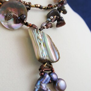 May include: A multi-strand necklace with brown beads, iridescent shell pendants, and purple glass beads. The necklace features a large iridescent shell pendant with a smaller shell pendant hanging from it.