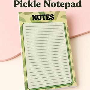 May include: A rectangular notepad featuring a light green background with a repeating dark green pickle pattern. The top of the page displays the word NOTES in bold black letters above a lined writing area. The notepad contains 50 tear-away pages.