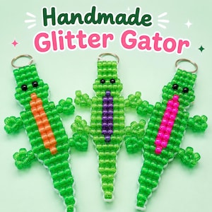 May include: Three handmade alligator keychains crafted from green glitter beads. Each features black bead eyes and a central stripe in orange, purple, or pink. A silver metal keyring is attached to the top of each. The text Handmade Glitter Gator is visible.