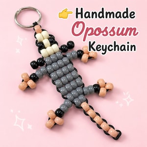 May include: A handmade opossum keychain crafted from grey, black, cream, and peach-colored plastic pony beads on a black cord with a silver metal ring. The text Handmade Opossum Keychain is visible in the upper right corner of the image.