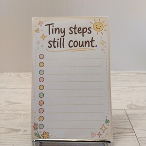 K&ouml;nnte beinhalten: Ein rechteckiger Notizblock mit dem Text Tiny steps still count in brauner Schrift. Das Design zeigt eine l&auml;chelnde Sonne, bunte Sterne, Herzen und eine Blume. Eine vertikale Spalte mit zehn runden K&auml;stchen befindet sich links neben dem linierten Papier.