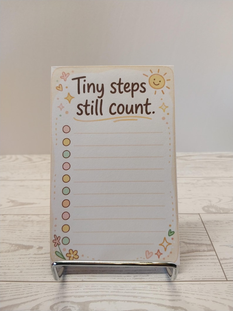 K&ouml;nnte beinhalten: Ein rechteckiger Notizblock mit dem Text Tiny steps still count in brauner Schrift. Das Design zeigt eine l&auml;chelnde Sonne, bunte Sterne, Herzen und eine Blume. Eine vertikale Spalte mit zehn runden K&auml;stchen befindet sich links neben dem linierten Papier.
