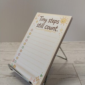 K&ouml;nnte beinhalten: Ein rechteckiger Notizblock auf einem silbernen Metallst&auml;nder mit dem Text Tiny steps still count. Das Papier hat einen cremefarbenen Hintergrund mit einer gelben Sonne, bunten runden K&auml;stchen und kleinen Blumen- und Herzillustrationen.