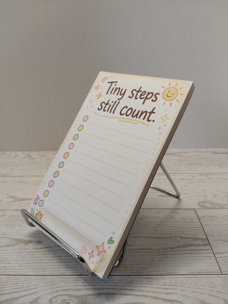K&ouml;nnte beinhalten: Ein rechteckiger Notizblock auf einem silbernen Metallst&auml;nder mit dem Text Tiny steps still count. Das Papier hat einen cremefarbenen Hintergrund mit einer gelben Sonne, bunten runden K&auml;stchen und kleinen Blumen- und Herzillustrationen.