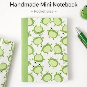 May include: A handmade mini notebook with a green spine and a white cover patterned with light green cartoon frogs. The text "Handmade Mini Notebook - Pocket Size -" is at the top of the image.