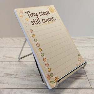 K&ouml;nnte beinhalten: Ein rechteckiger Notizblock auf einem silbernen Metallst&auml;nder. Das Papier hat einen cremefarbenen Hintergrund mit dem braunen Text "Tiny steps still count." und einem Sonnensymbol. Es enth&auml;lt eine vertikale Spalte mit farbigen Kreisen.