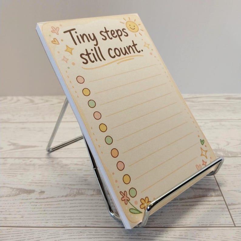 K&ouml;nnte beinhalten: Ein rechteckiger Notizblock auf einem silbernen Metallst&auml;nder. Das Papier hat einen cremefarbenen Hintergrund mit dem braunen Text "Tiny steps still count." und einem Sonnensymbol. Es enth&auml;lt eine vertikale Spalte mit farbigen Kreisen.