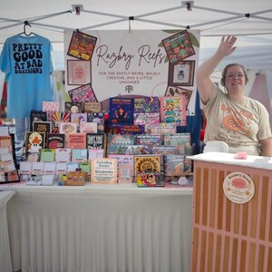 K&ouml;nnte beinhalten: Ein Marktstand mit dem Branding von Razbry Reefs, einem hellblauen T-Shirt mit dem Text Pretty Good At Bad Decisions und einer Auswahl an bunten Malb&uuml;chern und Notizheften. Ein Schild tr&auml;gt die Aufschrift For The Softly Weird, Wildly Creative, And A Little Unhinged.