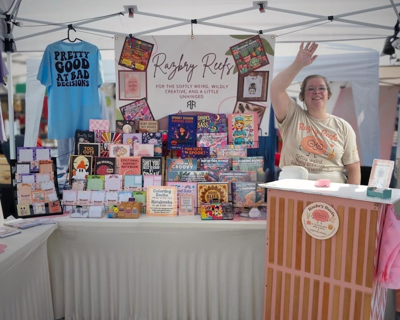 K&ouml;nnte beinhalten: Ein Marktstand mit dem Branding von Razbry Reefs, einem hellblauen T-Shirt mit dem Text Pretty Good At Bad Decisions und einer Auswahl an bunten Malb&uuml;chern und Notizheften. Ein Schild tr&auml;gt die Aufschrift For The Softly Weird, Wildly Creative, And A Little Unhinged.