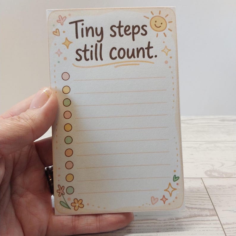 K&ouml;nnte beinhalten: Ein rechteckiger Notizblock mit cremefarbenem Hintergrund und dem braunen Text Tiny steps still count. Das Design zeigt eine Sonne, Sterne, Herzen und eine Blume sowie eine vertikale Spalte mit zehn bunten, runden K&auml;stchen neben linierten Zeilen.