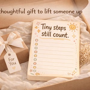 K&ouml;nnte beinhalten: Eine Pappschachtel mit einem Notizblock mit dem Text Tiny steps still count, einem kleinen braunen Geschenkpaket mit wei&szlig;er Schleife und einem Anh&auml;nger mit der Aufschrift For You sowie getrockneten Blumen. Der Deckel tr&auml;gt den Text A thoughtful gift to lift someone up.
