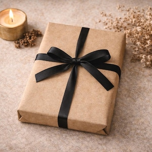 K&ouml;nnte beinhalten: Ein rechteckiges Geschenkpaket, eingewickelt in schlichtes braunes Kraftpapier und mit einem glatten, schwarzen Satinband zu einer mittigen Schleife gebunden. Das Paket liegt neben einer kleinen, brennenden goldfarbenen Kerze und getrockneten beigen Blumen.
