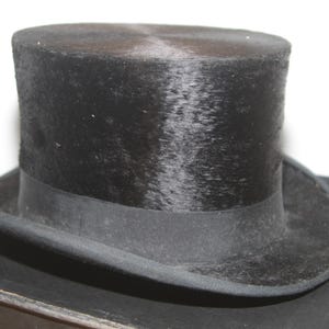 Puede incluir: Un sombrero de copa negro con una corona redondeada y un ala ancha. El sombrero tiene una cinta negra alrededor de la base de la corona. El sombrero está sobre una superficie negra.