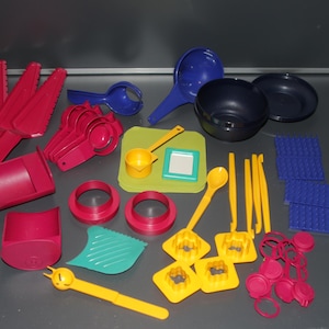 Op de afbeelding: Een verzameling kleurrijk keukengerei, waaronder rode spatels, maatbekers en lepels. Er zijn ook blauwe pollepels, kommen en snijplanken, samen met gele koekjessnijders en andere accessoires. De gereedschappen zijn gemaakt van plastic.
