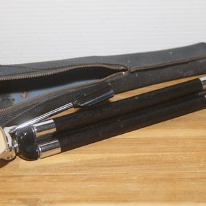 Op de afbeelding: Een vintage zwart camerastatief met zilveren accenten, gedeeltelijk uitgeschoven, naast de zwarte draagtas. De statiefpoten zijn van donker hout met zilveren uiteinden. De tas is open en toont de kop van het statief en een metalen verstelgereedschap.