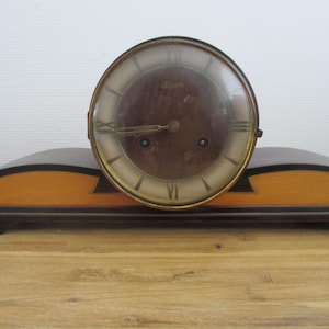 May include: A vintage wooden mantel clock with a circular face. The clock features a brown face with gold hands and Roman numerals. The base is a mix of light and dark wood, with a curved design. The clock is likely from the mid-20th century.