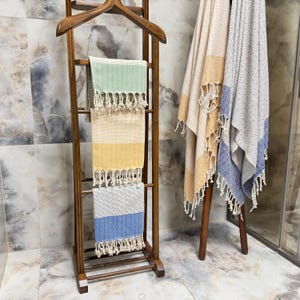 May include: Wooden towel rack with three colorful striped towels. The towels are green, yellow, and blue with white fringe. Two additional towels with fringe are on a separate wooden stand. The towels are beige, yellow, and blue.
