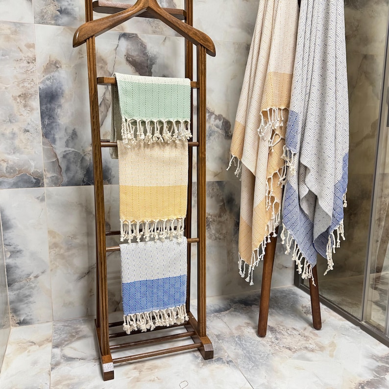 May include: Wooden towel rack with three colorful striped towels. The towels are green, yellow, and blue with white fringe. Two additional towels with fringe are on a separate wooden stand. The towels are beige, yellow, and blue.