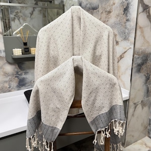 May include: A beige and white patterned Turkish towel with a geometric border and fringe hangs on a wooden chair. The towel is made of a soft, woven material and is displayed in a bathroom setting.