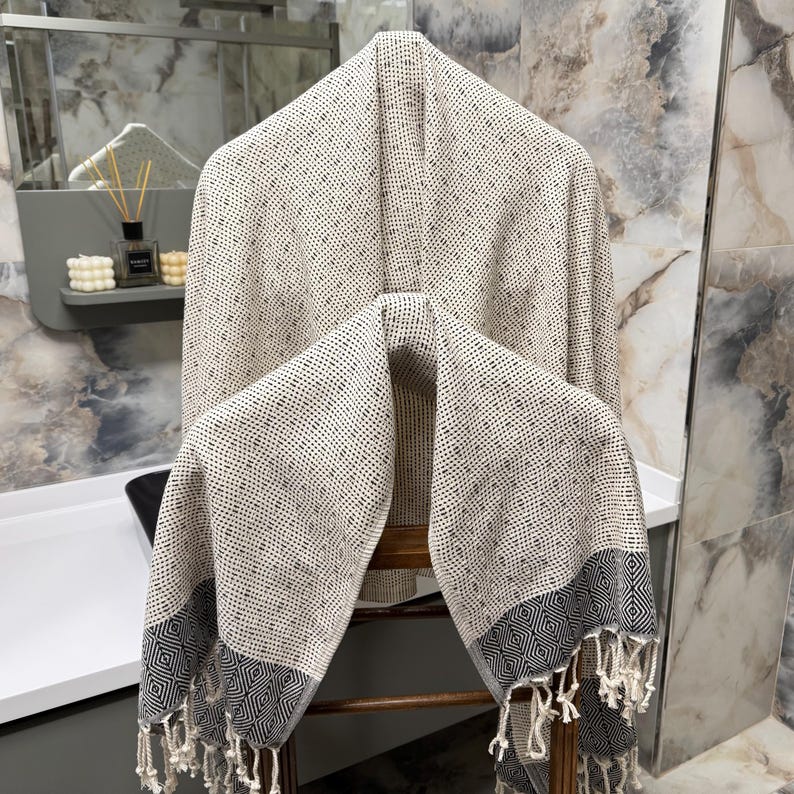 May include: A beige and white patterned Turkish towel with a geometric border and fringe hangs on a wooden chair. The towel is made of a soft, woven material and is displayed in a bathroom setting.