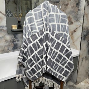 May include: A gray and white geometric patterned throw blanket with fringe. The blanket is draped over a wooden chair. The pattern is a grid of white lines on a gray background. The fringe is white.