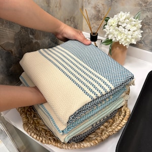 以下が含まれることがあります： クリーム色と青色の織り綿タオルが折りたたまれて積み重ねられています。タオルはストライプのデザインで、織り目が特徴です。タオルは編まれたバスケットに入っており、バスルームやスパの環境を示唆しています。