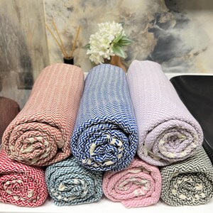 May include: Six rolled up towels in various colors, including pink, blue, red, and gray. The towels are made of a soft, woven fabric with a herringbone pattern.