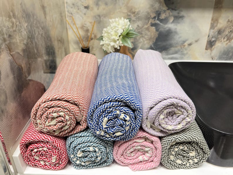 May include: Six rolled up towels in various colors, including pink, blue, red, and gray. The towels are made of a soft, woven fabric with a herringbone pattern.